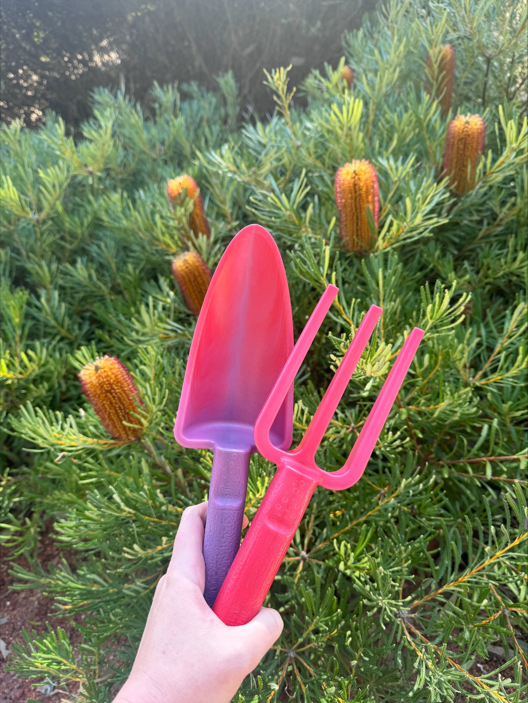 Pink gardening tools held in front of a green bush with red flowers.