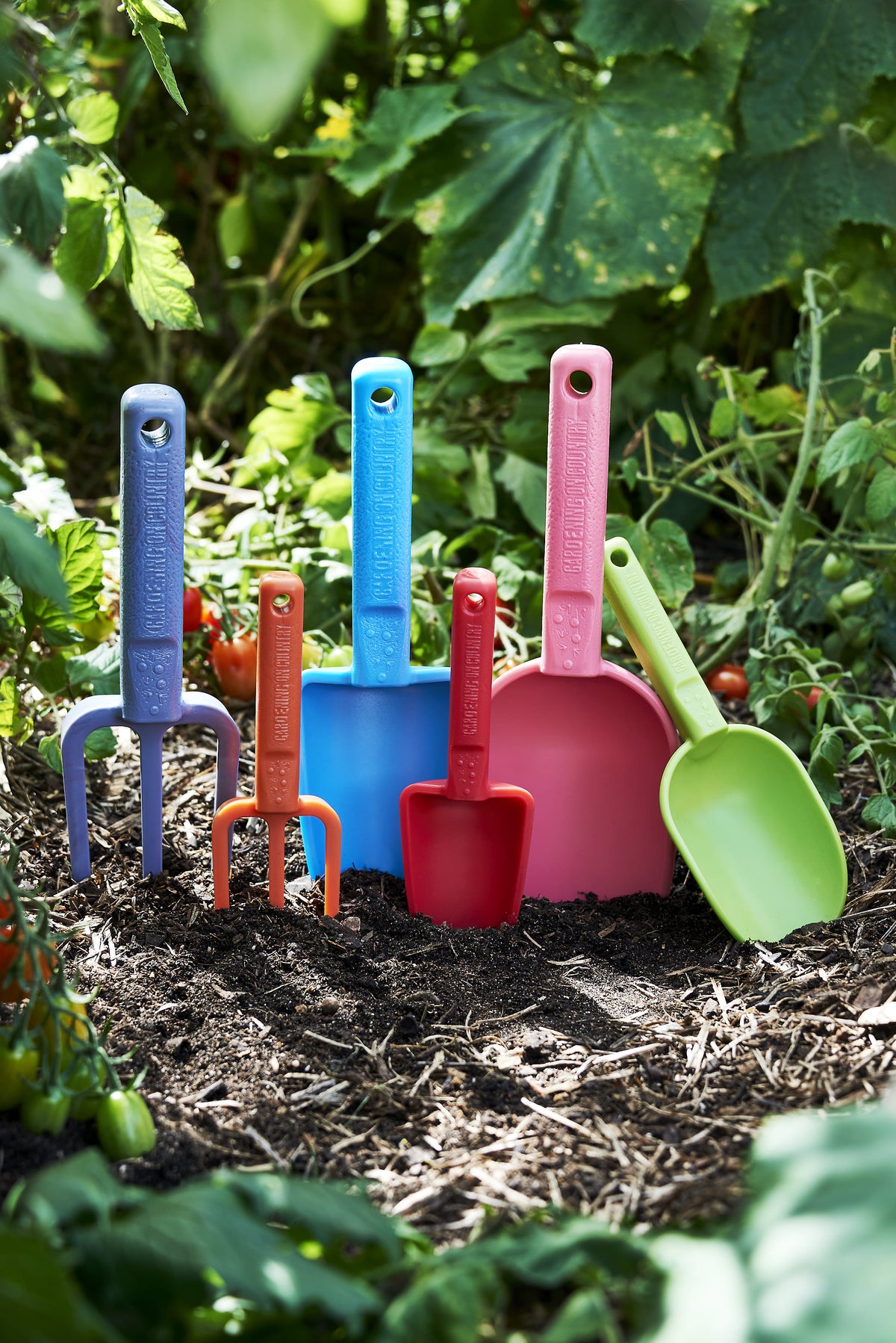 Set of colorful gardening tools including shovels and a fork on a garden bed with plants and tomatoes.