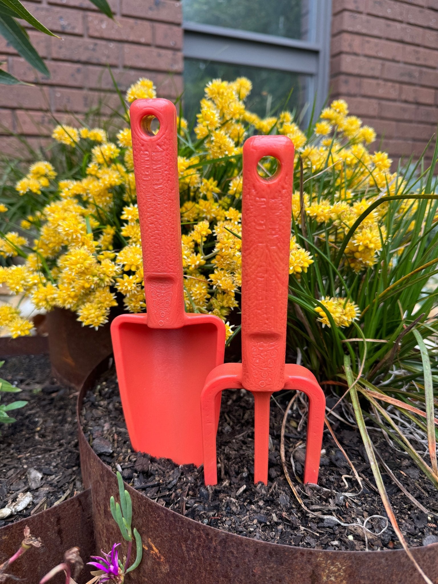 Red gardening tools (shovel and fork) in a garden bed with yellow flowers and a brick wall in the background.