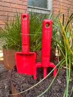 Red gardening tools, including a fork and trowel, in a garden setting with plants and soil.