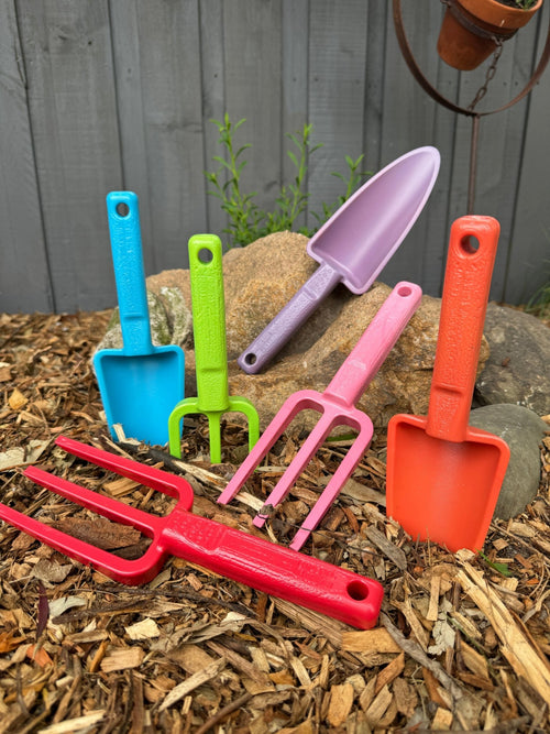 Set of colorful gardening tools including shovels and a fork on wood chips with a wooden fence background.