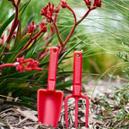 Two red recycled plastic gardening tools in a natural bush setting with plants and leaves.
