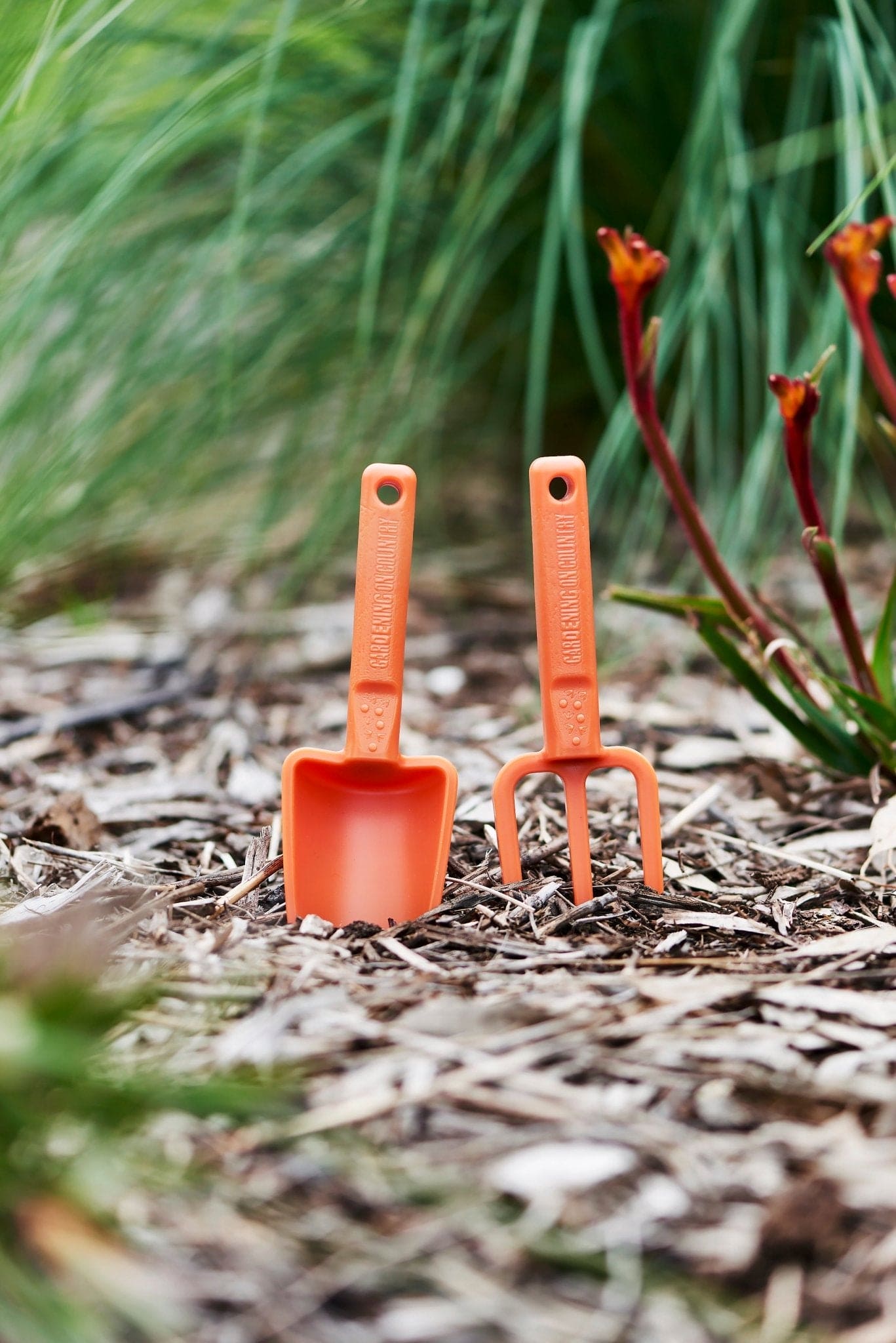 Orange-recycled-plastic-hand-trowel-and-hand-fork-kids-sitting-in-dirt-in-garden