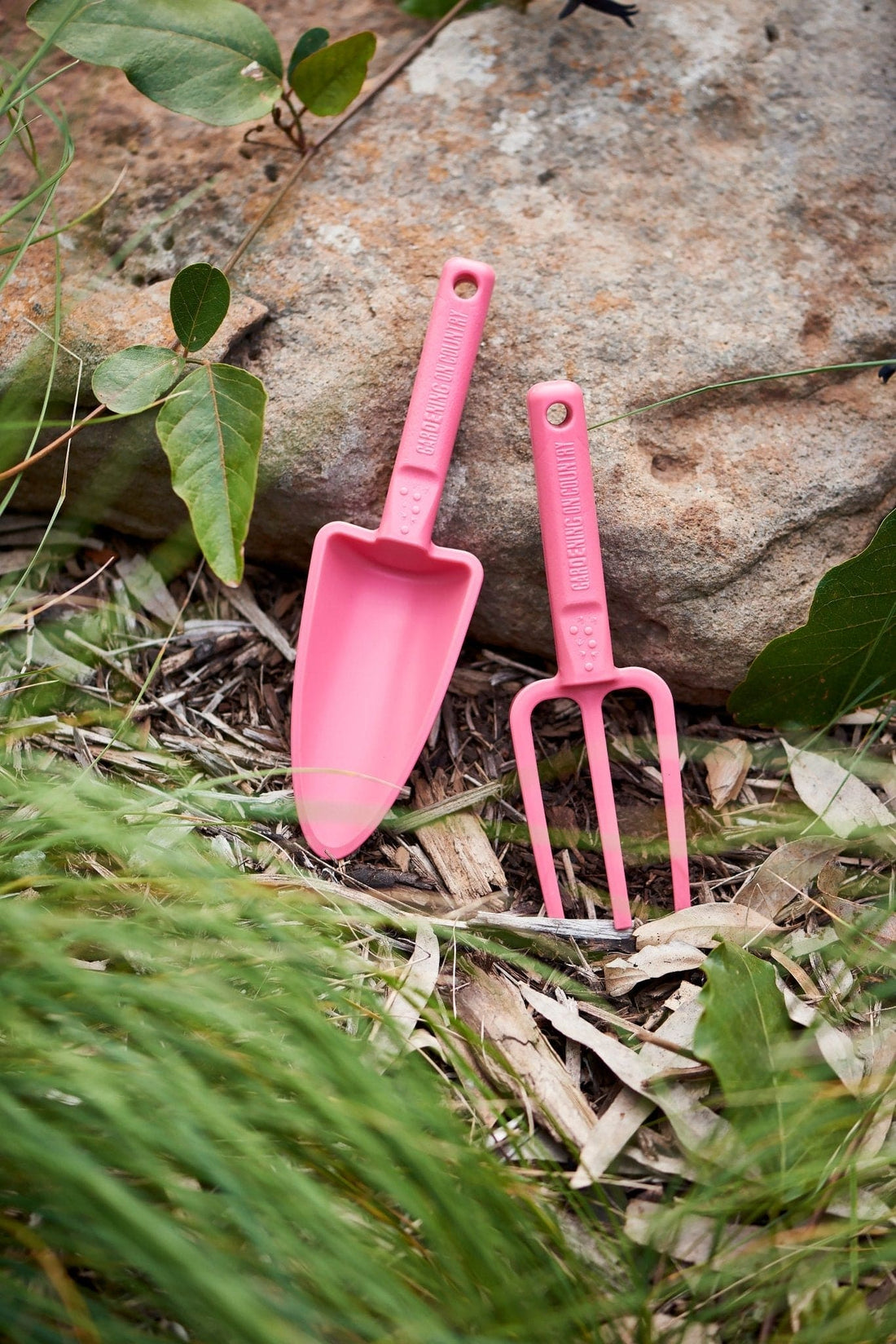 Pink-recycled-plastic-hand-trowel-and-hand-fork-kids-set-laying-against-rock-in-garden