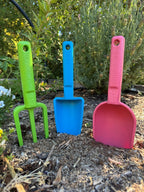 Three colorful garden tools (fork, scoop, and spade) on a garden bed with plants in the background. Pink, blue and green coloured eco garden tools.