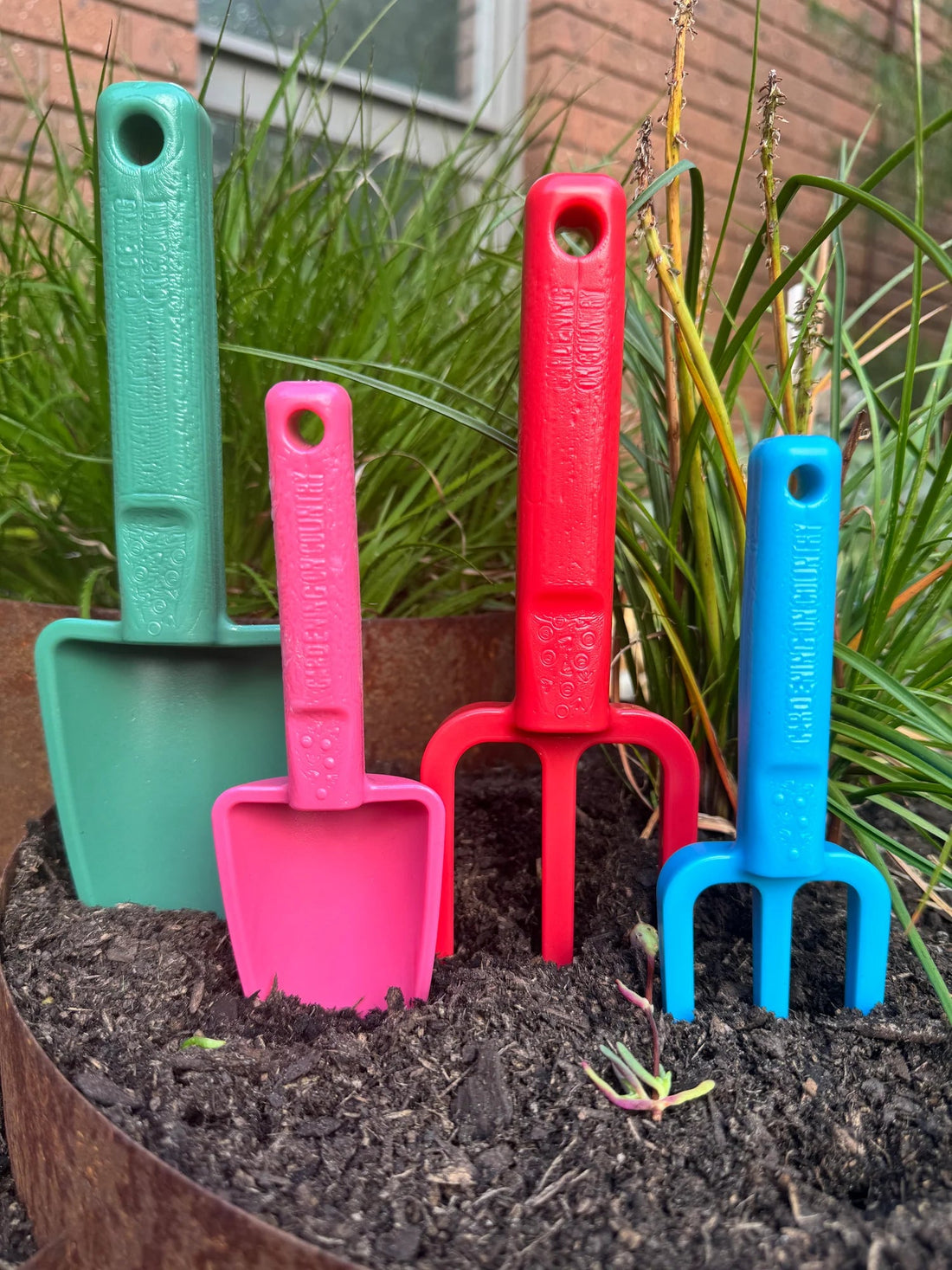 Set of colorful gardening tools including shovels and a fork on wood chips with a wooden fence background.