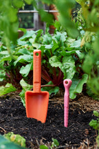 Recycled plastic orange and pink garden dibbler and trowel tools in a garden setting with plants and soil.