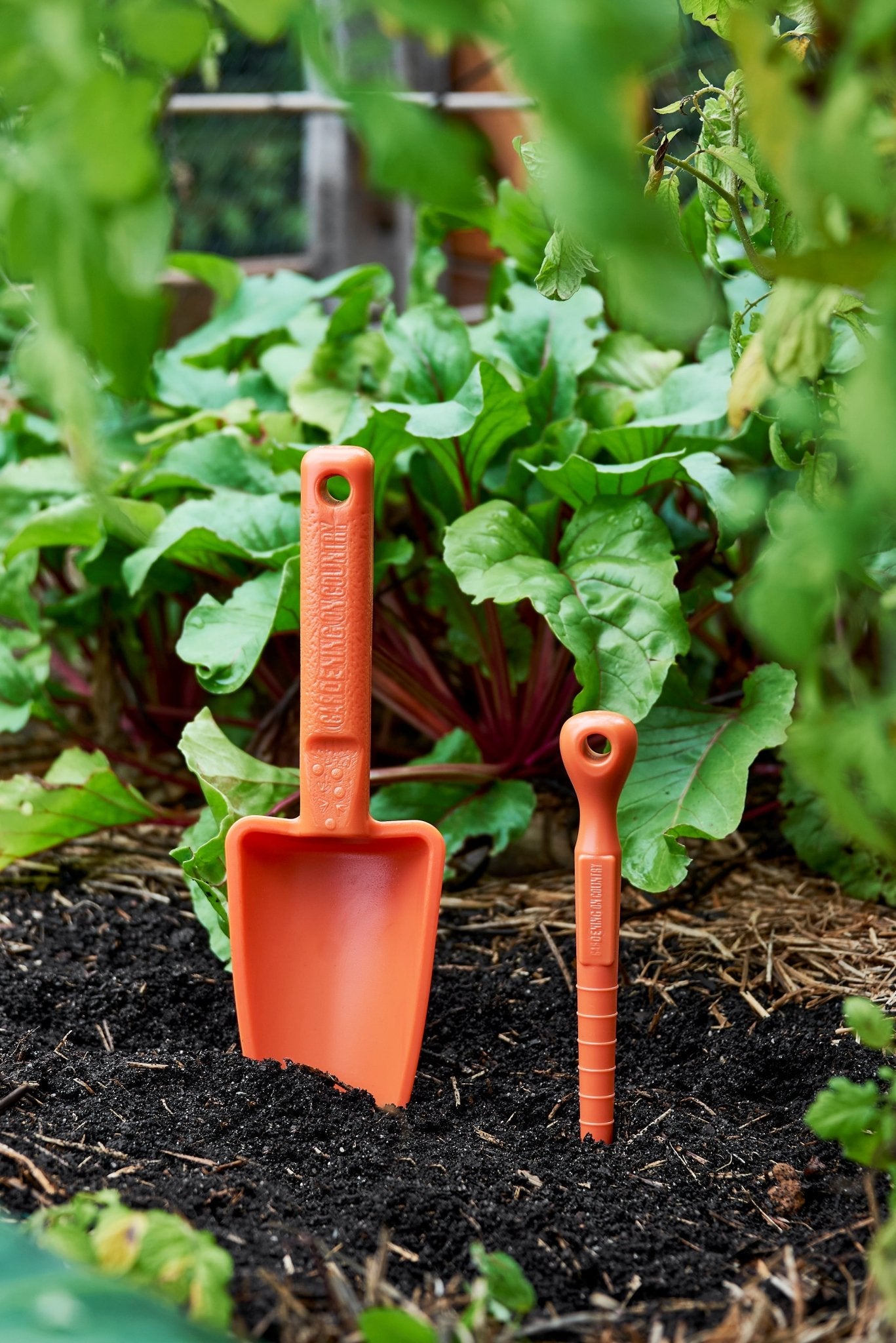 Two orange garden tools a dibbler and shovel in a garden setting with plants and soil.