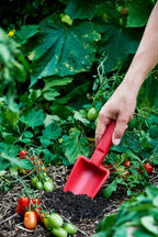 Hand using a red recycled plastic garden tool trowel in a garden with plants and vegetables.