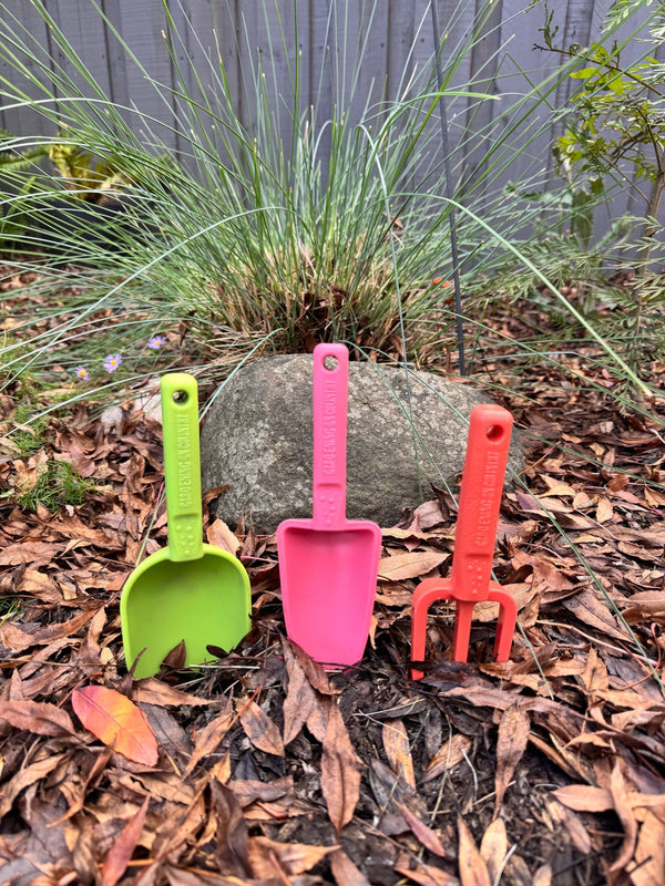 Three colorful recycled plastic garden tools (green, pink, red) on a bed of leaves and small rocks.