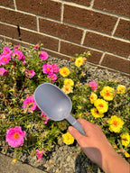 Purple recycled plastic gardening tool in front of pink and yellow flowers against a brick wall.