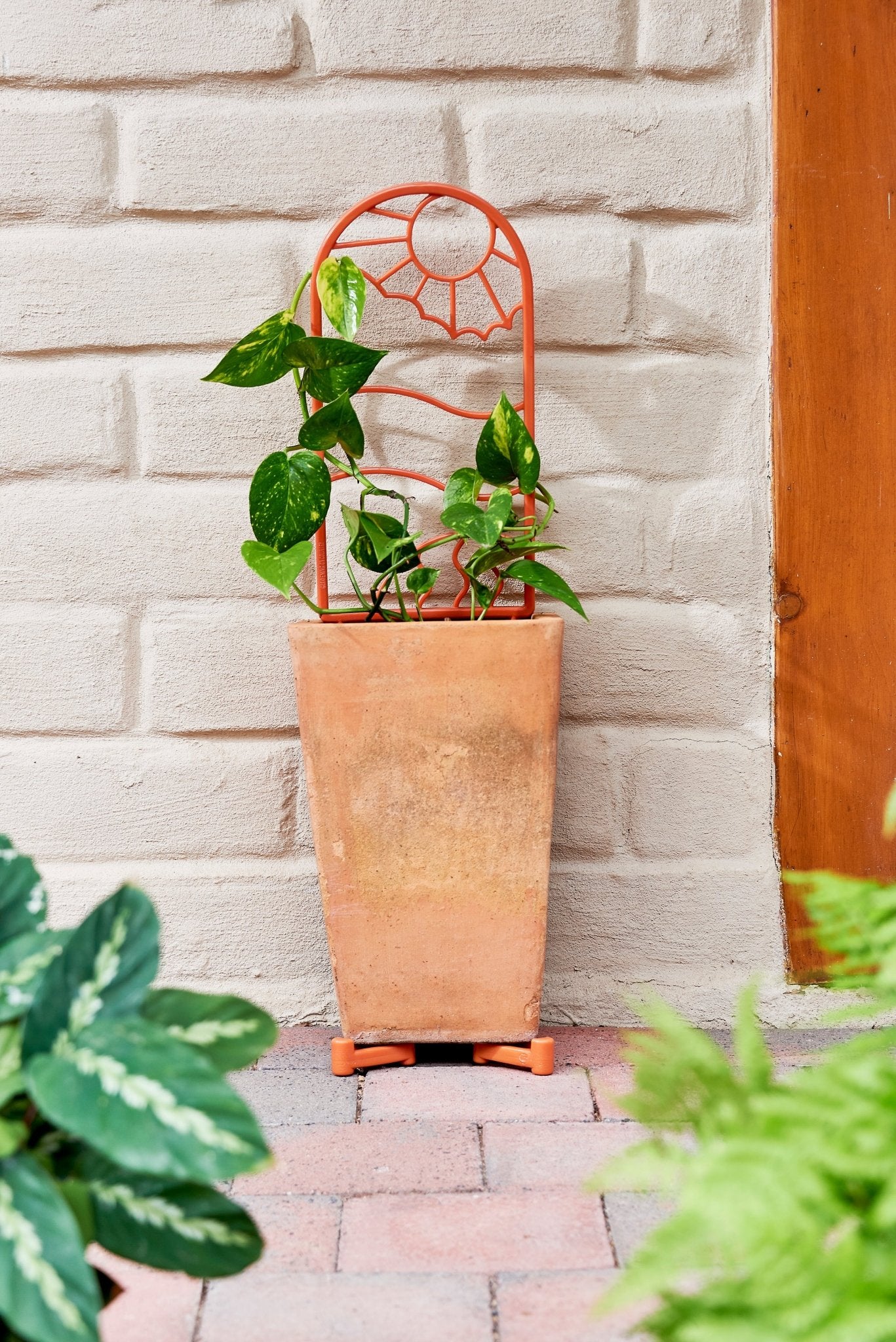 Terracotta planter with a decorative recycled plastic trellis and recycled plastic pot feet against a light brick wall.